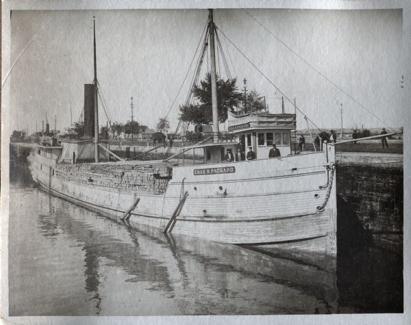 View across water towards the lumber steamer <i>Chas. B. Packard</i> docked with a load of lumber. Several crew members are standing on the deck, and men are standing on the walk behind the steamer. A railroad bridge is in the far background.