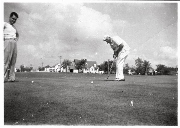 Carson Gulley putting on the ninth hole at the Westmorland Golf Course (1929-1944) in the 4200 block of Mineral Point Road as another man looks on. Several houses along the 400 block of Holly Avenue and the 4100 block of Euclid Avenue can be seen in the background.