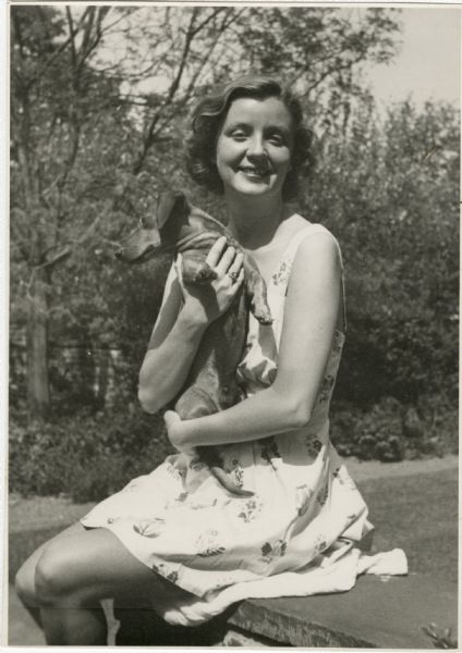 Mary Griggs Burke as a young woman, sitting outdoors at Forest Lodge smiling and holding a dachshund dog. 
