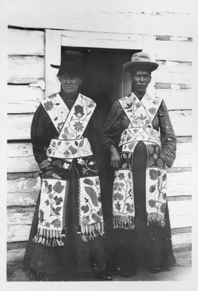 Two Ojibwa women in the Mille Lacs and Rainy Lake standing in front of a doorway. They are wearing traditional Indian attire, including beaded bandolier bags.