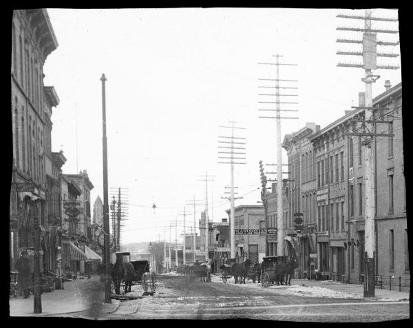 View down East Main Street, with a number of horse-drawn carriages parked at the curbs. Snow is in the street. There is a barber's pole on the sidewalk on the left, and a sign above a storefront on the right reads: "Madisonian Book and Job Printing."