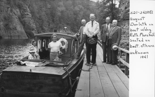 August Derleth posing on a boat tied up to a pier on the Wisconsin River. With him are Ruth Marshall (seated), another woman, and a dog. Four men are standing on the pier next to the boat.