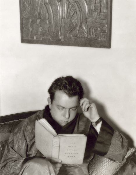Director Lewis Milestone reads the novel "All Quiet on the Western Front" while sitting on a sofa. Milestone directed the 1930 film version of the novel.