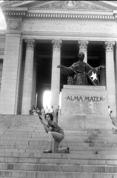 A woman posing on the steps of the University of Havana in Cuba in front of a statue. She is kneeling and holding a rifle. The statue is draped with a cloth or flag, and on the base are the words: "Alma Mater." In the background at the top of the steps is a group of people watching. A sign carved above the columns on the building reads: "Universidad de la Habana."
