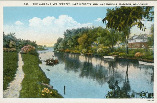 Elevated view from shoreline of the Yahara River, with a path running along the shoreline on the left, and a bridge in the distance. Boats are docked near several small piers on both sides of the river. A large house is on the far shoreline on the right. Caption reads: "The Yahara River Between Lake Mendota and Lake Monona, Madison, Wisconsin."