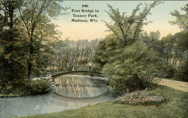 View from lawn towards the arched foot bridge and drive at Tenney Park. A young man is standing on the bridge. Caption reads: "Foot Bridge in Tenney Park, Madison, Wis."
