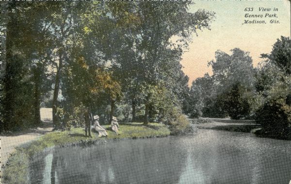 View along shoreline towards three people relaxing on the bank of the Tenney Park lagoon. A man is standing near two women who are wearing long dresses and large hats. There is a path and a glimpse of a bridge behind them on the left. Caption reads: "View in Tenney Park, Madison, Wis."