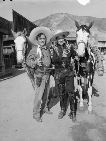 Publicity photo of the Cisco Kid (Duncan Renaldo) and Pancho (Leo Carillo) standing next to their horses. They are both holding and pointing guns.