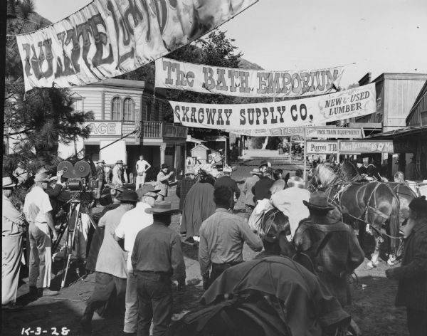 The cast and crew of the TV show "Klondike" on an outdoor set designed like the main street of a western town. There is a camera operator on the left. Banners are hanging above the street.