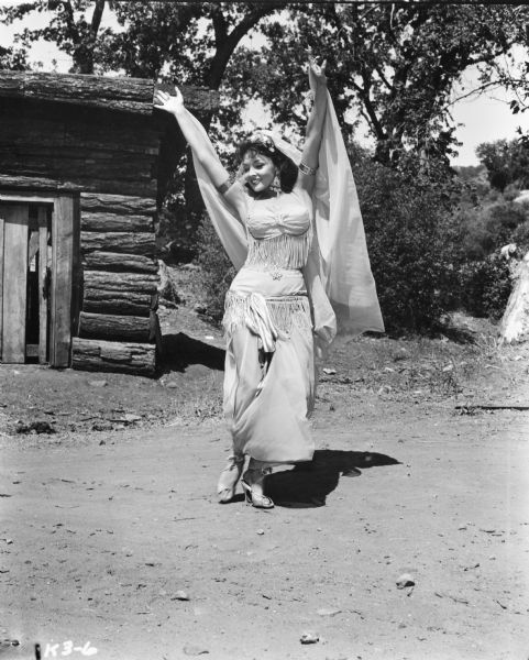 A belly dancer performing outdoors on the set of the TV show "Klondike." She has her arms raised above her head holding a large scarf. There is a log cabin behind her.
