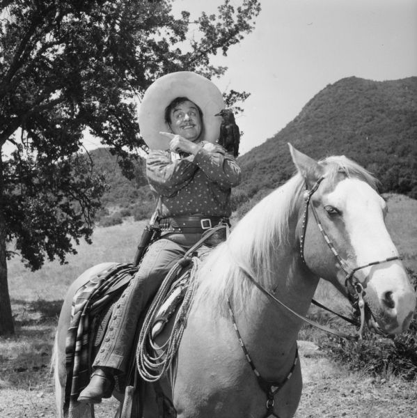 Outdoor publicity still of Pancho, actor Leo Carillo, sitting on horseback. He is posing with his arms crossed in front of his chest, and a bird (stuffed?) is perched on his shoulder.