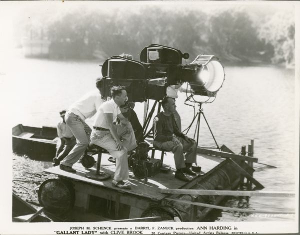 The director and four other men sitting and standing on a raft holding the motion picture camera while shooting the film "Gallant Lady." There are two cameras and a large light. The raft is partially on the shoreline of the lake, and a man is sitting on the side of a boat behind the raft.