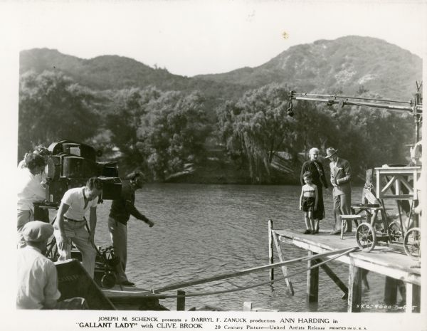 The cast and crew of the film "Gallant Lady" preparing to shoot a scene on a lake. The motion picture camera and crew are on a raft on the left, and are shooting a man, woman and boy who are standing on a pier on the right. Mountains and trees are on the far shoreline in the background.