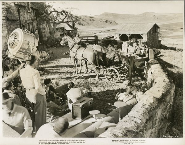 Elevated view of the set of the film "Wuthering Heights." The crew is standing and sitting behind the camera which is on the ground. An actor and actress are sitting in a buggy pulled by a team of two horses.