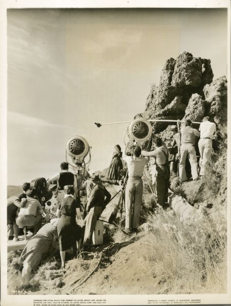 The crew on the film "Wuthering Heights" shooting a scene with an actress on a cliff. The camera is on the left with two large lights, and two men holding the microphone boom in the cente.