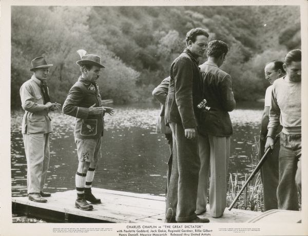 Charlie Chaplin, dressed in a German ethnic costume, standing on a pier on a lake with crew members during the filming of "The Great Dictator."