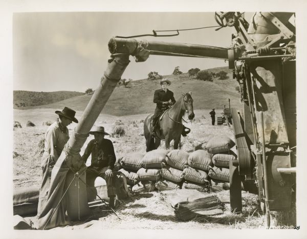 Production still from the film "Of Mice and Men." A man is sitting on a horse while two other men are sitting and standing next to a harvesting machine in a field.