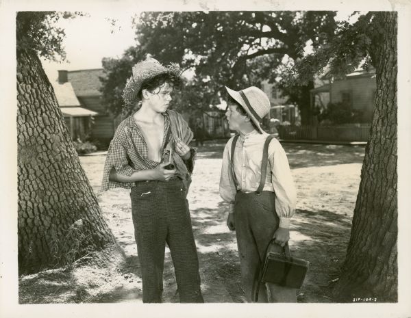 Tom Sawyer (Tommy Kelly) looking at Huckleberry Finn (Jackie Moran) while standing outdoors. Tom is holding books on a strap, and Huck is holding a pipe.