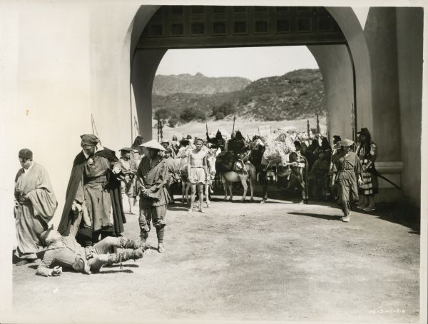 Cast members of the film "The Adventures of Marco Polo" standing in the entryway of a fortress. Marco Polo (Gary Cooper) is holding the hand of Binguccio (Ernest Truex) who is laying on the ground in the foreground.