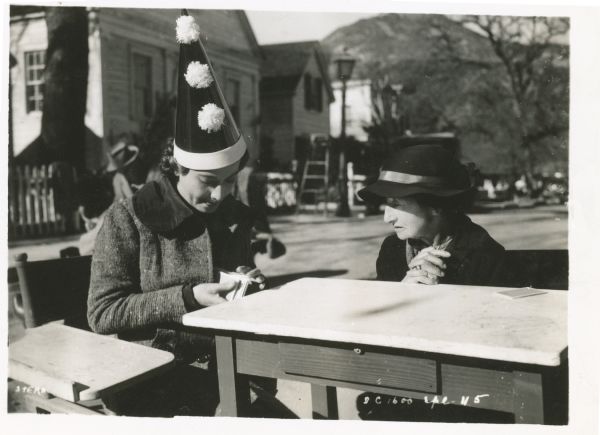 Merle Oberon sitting next to an older woman at a table outdoors. Oberon is wearing a pointed clown hat with three pompoms on it. She is holding cards in her hand.