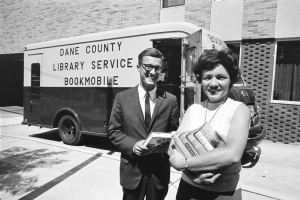 Dane County Library Service Director, Donald Lamb and Librarian Barbara Larson standing outdoors in front of the Bookmobile parked at the Madison Public Library on the corner of West Mifflin and North Henry Streets. Both of them are holding books.
