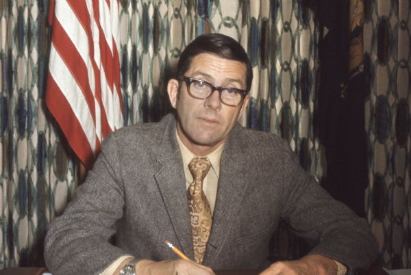 Michael Early was State Assembly Representative for Buffalo, Pepin, and Pierce counties, and a Democrat. He is seated and holding a pencil, with the American and Wisconsin state flags behind him.