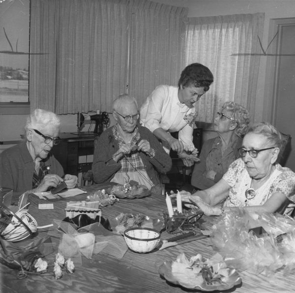 Four residents of the Barron County Nursing Home in Cumberland are making crafts around a table. A caption on the photograph reads: "This group in occupational therapy is making items of handicraft which are placed on sale in the nursing home lobby. Proceeds go to buy more materials for use by the some 20 residents who 'attend classes' twice a week." From right are Mrs. Crisler, Rice Lake, Gurine Anderson, Cumberland, Edna Rust, occupational therapist, Lois Herning, Rice Lake, and Alice Erwin, Barron.