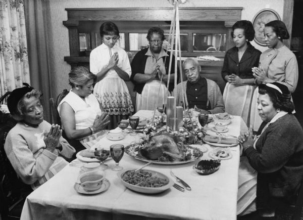 Group portrait of several people sitting or standing around a table with a meal displayed on it. All the women, and one man, are folding their hands as if in prayer. Caption reads: "A family style Christmas dinner was enjoyed by four residents of the Community Home for the Aged and Convalescent Wednesday at the home of Mrs. Mamie W. Lewis, 3244 N. 14th st. Gathered around the table are (from left) Mrs. Frankie Thompson, 80, a guest; Mrs. Mattie Carter, 59, guest; Mrs. Lewis; Mrs. Lillie Graham, who cooked the dinner; Hayes Harris, 74, guest; Mrs. Betty Rowe and Mrs. Leroy Mitcham, assistant hostesses, and Mrs. Estella Ford, 48, guest. Mrs. Lewis arranged the party to give some of the residents of the home a change from everyday routine."