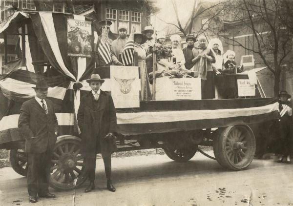 Parade float on which ten women are standing, some posing with knitting needles or behind a sewing machine. Two men and one woman are standing on the ground next to the float. Signs on the float identify the National League for Women's Service, a constructive patriotism organization for women. One sign reads: National League for Womens Service has pledged to knit 4000 garments for the Red Cross.