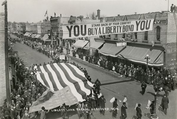 Elevated view of a parade, including a large American flag carried by several men. Spectators line the streets and some are standing on rooftops. A banner above the street reads: "YOU Stand back of your country / Buy a Liberty Bond YOU."
