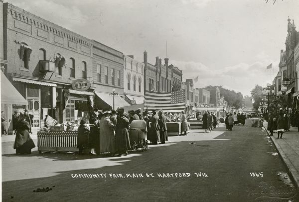 View down Main Street towards women looking at tables set up along the business district. The tables have been decorated with American flags. Shop signs for La Palina cigar store and Schaller's Bakery on the commercial building that line the street.