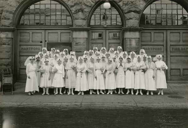 Outdoor group portrait of Red Cross nurses standing in front of the Hartford Fire Department building for Tag Day, which was a fundraising event.