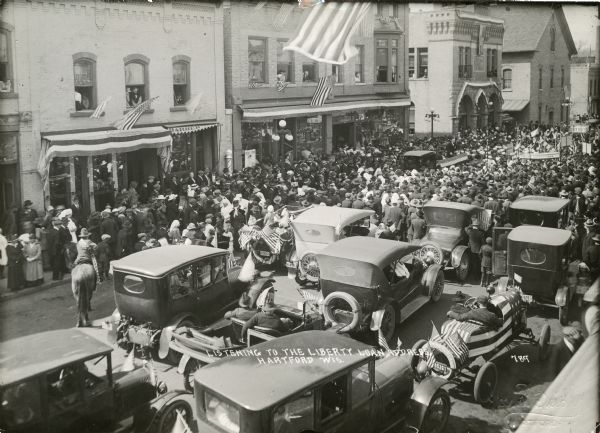 Elevated view of a crowd that is gathered to listen to a man speaking from the back of a truck. Several cars are in the foreground, as well as one uniformed person on a horse. People are also listening from windows above shops. Caption reads: "Listening To The Liberty Loan Address. Hartford Wis."