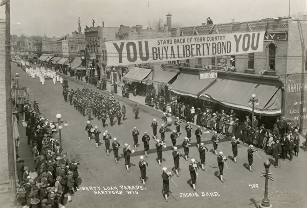 Elevated view of a marching band coming down a street playing instruments. They are followed by marching soldiers, who are followed by marching nurses. Bystanders flank the street. Overhead, a banner reads "YOU Stand Back of Your Country / Buy a Liberty Bond YOU."