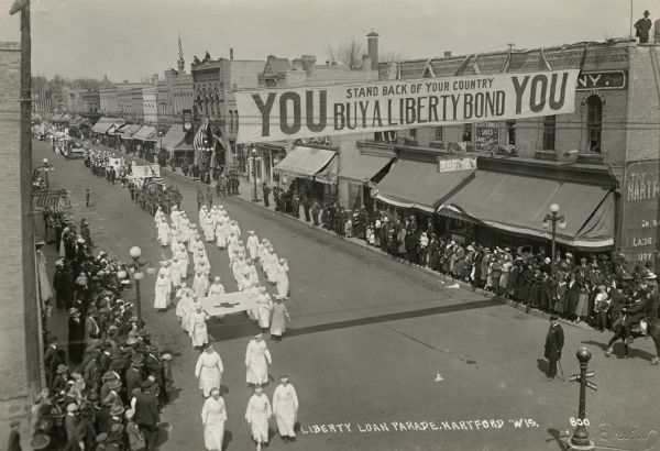 Elevated view of Red Cross nurses marching along a street, holding the Red Cross flag. Bystanders flank the street. Overhead, a banner reads "YOU Stand Back of Your Country / Buy a Liberty Bond YOU."