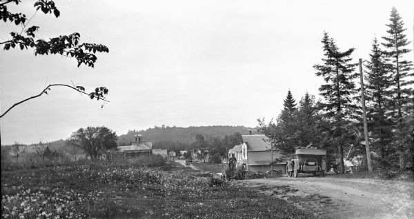 View from low hill of a man standing as he is driving a team and wagon up Egg Harbor Road, traveling north. An automobile is parked in the foreground along the road, and a second is seen in the distance. The 1871 Levi Thorp house, now called the "Cupola House" is on the left; there is a wood frame commercial building on the opposite side of the road. Cord wood is stacked alongside the building.