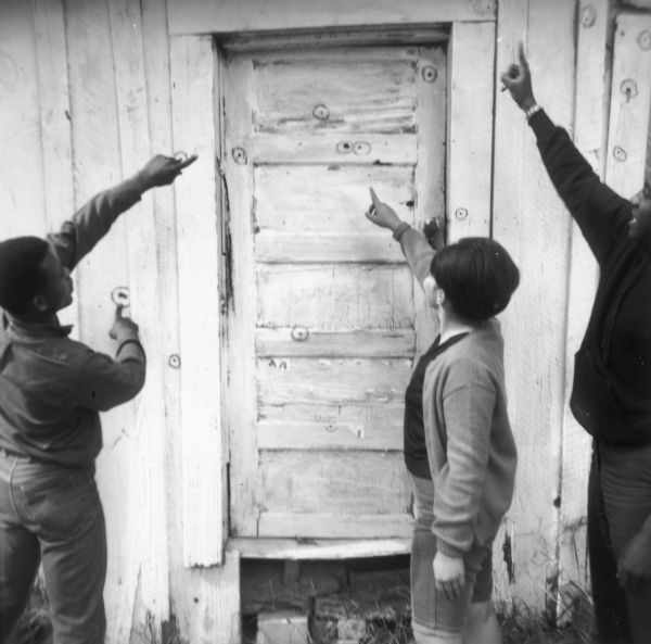 Three individuals point towards shotgun holes in a door and on the side of a building, highlighted by circles. The building is the Pheba Freedom House, used by civil rights volunteers. It was later burned down.