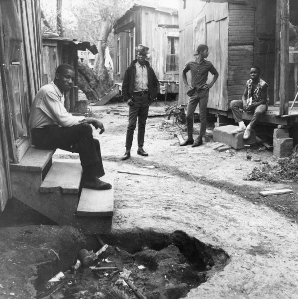 Four men gathered outdoors. Two sit on stoops of houses while two stand in the dirt pathway. More homes are visible in the background. There is a garbage or fire pit in the foreground.<p>Fifty-one people, twenty-five under the age of 16, living in 8 buildings share two toilets and 4 water hydrants in the South Wall Street neighborhood of Natchez.