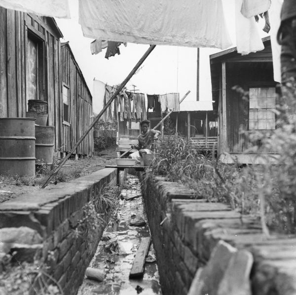 A girl fills a bucket at a water hydrant near a drainage ditch.  Buildings are on either side, and laundry hangs from clotheslines above.<p>“Row upon row of dilapidated buildings, three rows deep, characterize this slum. The city has no housing code and continues to ignore the slums of Natchez. No pavement, no streetlights, 10 outside flush toilets in 2 centrally located shanties shared by 114 people. The 125 persons living in this area share eight water faucets; of these 8, two are inside (installed by tenants) and 6 are outside.”