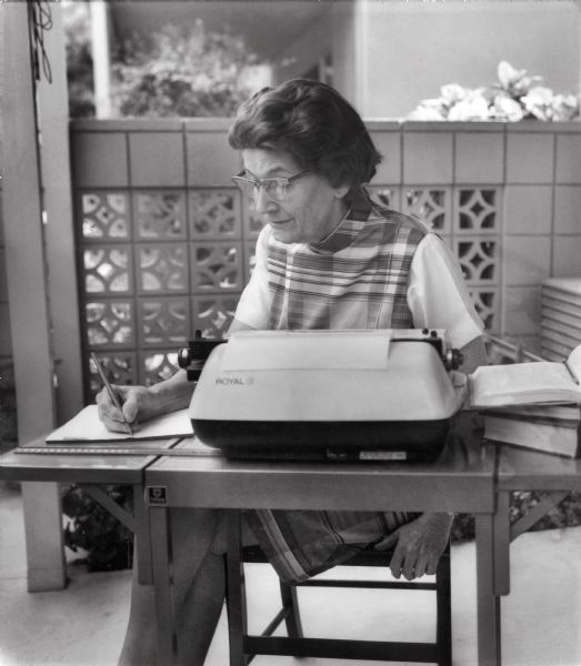 A candid portrait of Alice E. Smith sitting behind a typewriter. She appears to be sitting on an outdoor patio, and is writing on a pad of paper.