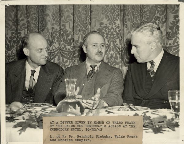 Seated at a table, from left to right, are Reinhold Niebuhr, Waldo Frank, and Charles Chaplin. They are at the Commodore Hotel at a dinner in honor of Frank, given by the Americans for Democratic Action. 