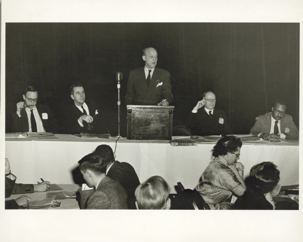 View from audience of Reinhold Niebuhr speaking from a podium at an Americans for Democratic Action event. He is holding a cigarette in his hand.