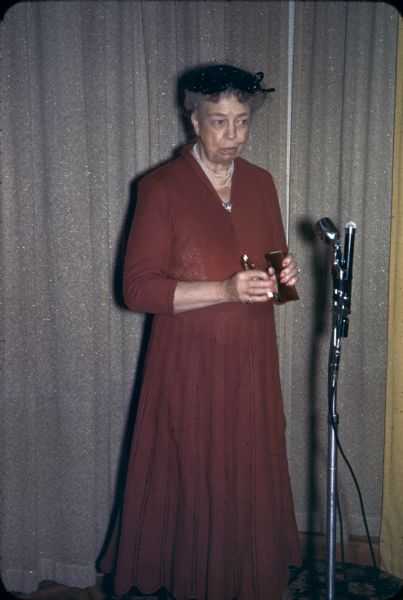 Eleanor Roosevelt standing in front of a microphone, speaking at a press conference. She is holding eyeglasses in her hand.