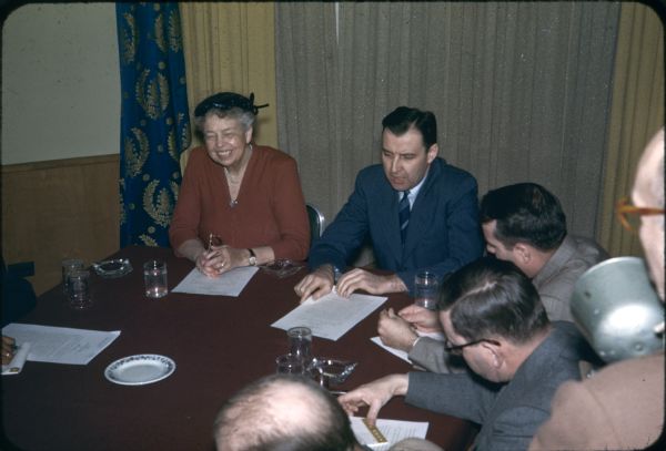 Eleanor Roosevelt, smiling and holding eyeglasses in her hands, is sitting at a table with several men during a press conference. 