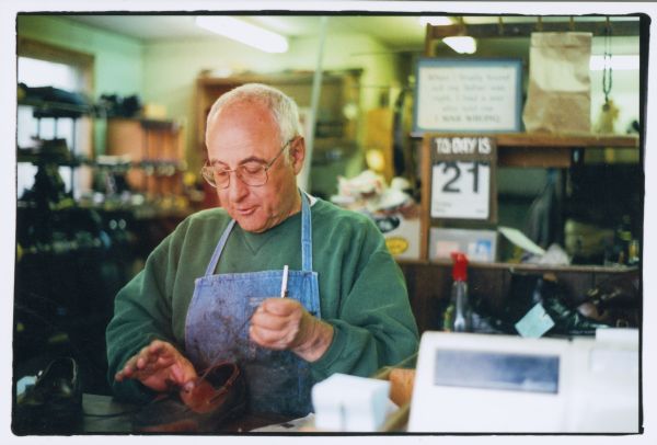 George Fabian at work at his business, Park Street Shoe Repair, at 609 South Park Street. He is wearing a denim apron and is lacing a shoe.