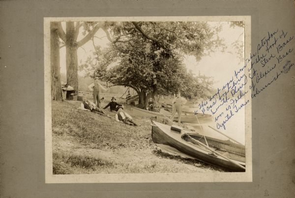 Several men relaxing on the grass at the edge of a lake. There are boats pulled up on the shore near them. The man standing on the far right with his hand on his hip is Alex Baas. Karl Fauerbach wrote this on the photograph in blue ink: "This boat was from the Stephen Baas boat livery at the foot of South Blair St. Steve Baas was is [sic] father of Alexius Baas, Capitol Times columnist, + music teacher."