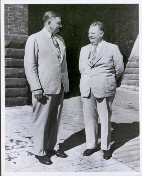 Karl H. Fauerbach and Don Huseby standing together in front of Fauerbach Brewing Company. They are wearing suits and smiling at one another. 