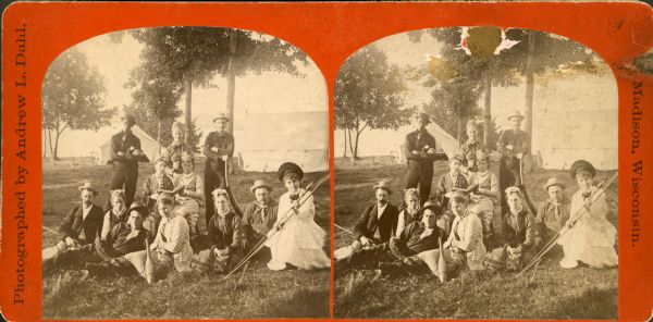 Stereograph of a group of people posing by the lakeshore of Maple Bluff with tents in the background. One of the woman is holding an archery bow, two women are sitting in chairs and holding a open book together, and a woman sitting on the grass has a bowling pin resting by her knee. A man standing in the back of the group is holding a long horn near his side.