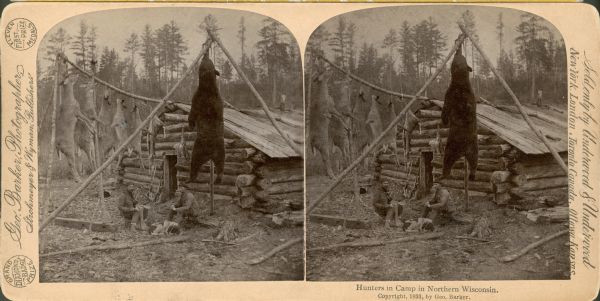 Two men are sitting outside of a cabin at a hunter's camp. A dog is curled up at their feet. A dead bear and several dead deer are hung up behind them on poles.