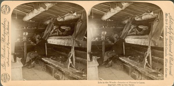 The interior of a rustic hunter's cabin. There is a rifle propped up on a bench in the foreground. A man is reclining in a bunk, while another man is sitting on a bench.